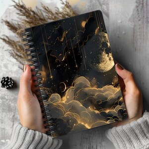 Op de afbeelding: Een spiraalgebonden notitieboek met een illustratie van een nachtelijke hemel op de kaft. Het ontwerp toont een grote, getextureerde gouden maan, verspreide sterren en gestileerde wolken met gloeiende gouden omlijningen op een zwarte achtergrond.