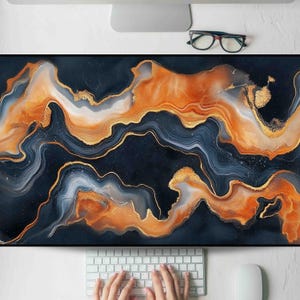 May include: A large, rectangular desk mat with an abstract design. The mat features flowing patterns in shades of navy blue, orange, gold, and white. The design resembles marble or agate, with a glossy finish. A keyboard, mouse, and glasses are visible.
