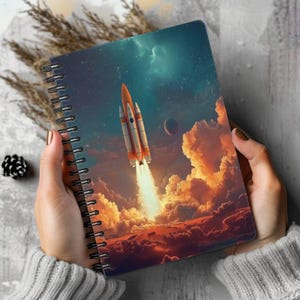 May include: A spiral-bound notebook featuring a graphic illustration of an orange and white rocket launching into a dark blue starry sky above bright orange clouds and a planet. The cover art depicts a space scene with a glowing exhaust plume.