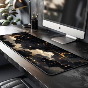 May include: A rectangular desk mat featuring a celestial design with a black background, gold accents, and cloud-like shapes. The mat has a leather-like border and is placed on a dark desk, next to a computer monitor.