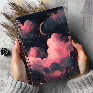 May include: A spiral-bound notebook featuring a dark night sky illustration with a crescent moon and large, voluminous clouds in shades of pink, magenta, and deep purple. The cover art depicts a celestial scene with scattered stars.