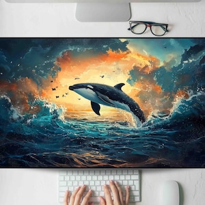 May include: A vibrant desktop mat featuring a breaching orca whale in a dynamic ocean scene. The artwork showcases a black and white whale against a backdrop of swirling blue waves and a fiery orange and yellow sky. Small birds are scattered throughout the scene.