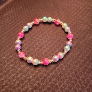 May include: A bracelet made of iridescent pink, blue, and white beads. The beads are strung on a clear elastic cord.
