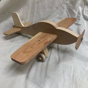 May include: A wooden toy airplane with a light brown finish. The plane has a propeller on the back and wheels on the bottom.
