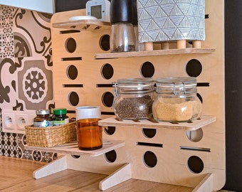 May include: A wooden shelf unit with multiple shelves and holes for storage. The shelves are filled with various items, including jars of spices, a digital clock, and a potted plant. The shelf unit is made of light wood and has a natural finish.