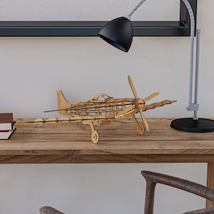 May include: A wooden model airplane with a brown finish sits on a wooden desk. The airplane has a propeller and is in flight position. There are books stacked on the desk and a lamp with a black shade.