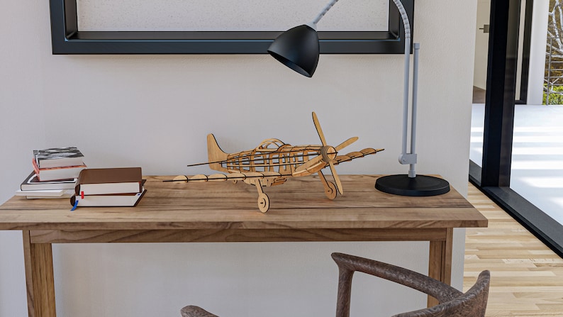 May include: A wooden model airplane with a brown finish sits on a wooden desk. The airplane has a propeller and is in flight position. There are books stacked on the desk and a lamp with a black shade.