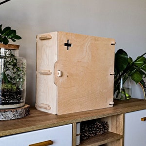 Puede incluir: Una caja de madera de color claro con un recorte en forma de cruz y varias pestañas de madera. La caja está sobre una estantería de madera con un terrario y plantas en el fondo. La caja parece ser un contenedor de almacenamiento.