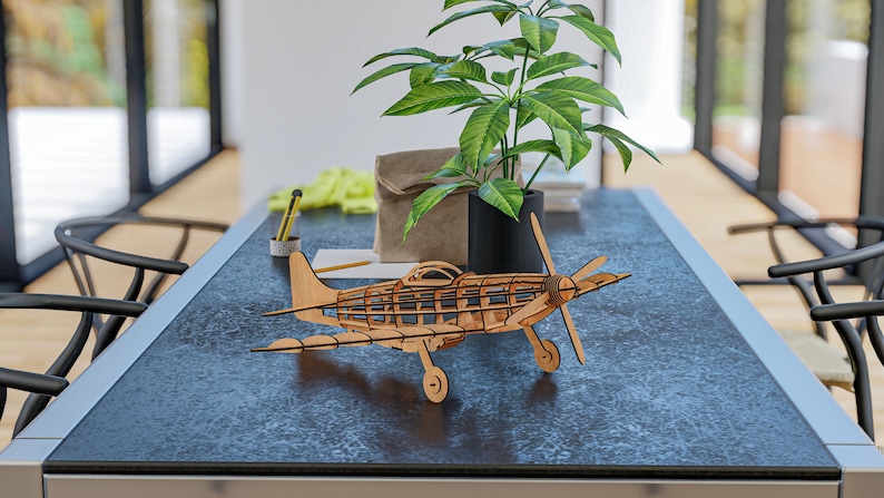 May include: A wooden model airplane with a brown finish sits on a dark gray table. The airplane has a propeller and wings. A potted plant with green leaves is in the background.