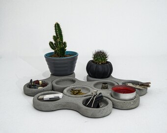Puede incluir: Un conjunto de seis platos de hormigón gris con bordes redondeados, dispuestos en forma circular. Los platos están llenos de varios objetos pequeños, incluyendo un pequeño cactus, una vela, una llave y una unidad USB.