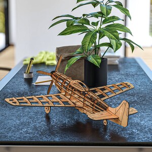 May include: A wooden model airplane with a detailed, open-frame design. The model is made of light brown wood and has a black base. The model is sitting on a dark gray table with a plant in a black pot in the background.
