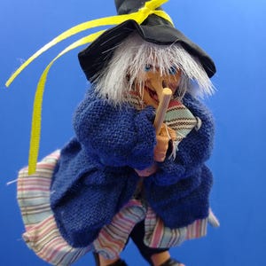 Puede incluir: Una muñeca de bruja caprichosa con un sombrero negro adornado con una cinta amarilla. La muñeca tiene cabello blanco, ojos azules y un bastón de madera. Viste un abrigo azul tejido y una falda a rayas, con sandalias negras. Una encantadora decoración de Halloween.