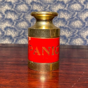 May include: A brass and red container with the word "PANIC" printed on the red section. The container has a gold-colored top and bottom, and it sits on a wooden surface.
