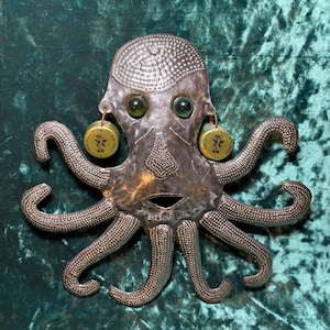 May include: A detailed silver octopus sculpture with green glass eyes and dangling earrings. The octopus has eight textured tentacles and a detailed head. The earrings are circular with text. The sculpture is set against a teal background.