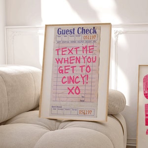 Puede incluir: Dos impresiones artísticas enmarcadas. Una presenta un recibo con el texto "TEXT ME WHEN YOU GET TO CINCY! XO" en rosa. La otra impresión muestra "CINCINNATI OH 10" en una fuente rosa pixelada. Ambas están en marcos de madera.