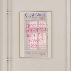 Może przedstawiać: Oprawiony wydruk w stylu vintage z rachunkiem dla gości. U góry napis "Guest Check", a różowym kolorem słowa "TEXT ME WHEN YOU GET TO WACO! XO". Wydruk jest w złotej ramie i wisi na białej ścianie.