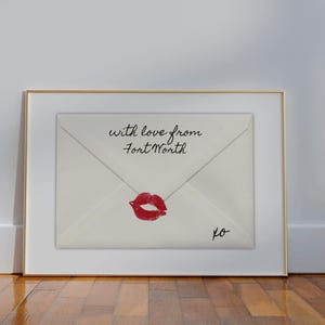 Op de afbeelding: Ingelijst kunstwerk met een crèmekleurige envelop met een rode lipstickkus en de tekst "with love from Fort Worth". Het kunstwerk staat in een gouden lijst, tegen een witte muur en een houten vloer.