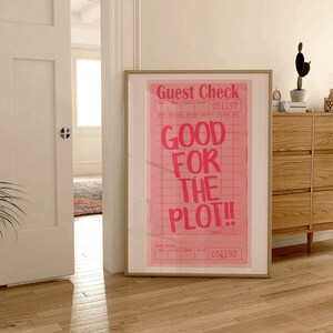 以下が含まれることがあります： 薄い木の床に置かれた、ピンクと白のポスター。ポスターには「Guest Check」と「Good for the Plot!」の文字が書かれている。部屋には木の引き出しと植物がある。