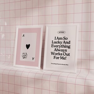 Puede incluir: Dos impresiones enmarcadas, una con fondo rosa y una carta de juego de as de corazones negra con el texto "I'm a lucky girl!" y la otra con fondo blanco y el texto "AFFIRM I Am So Lucky And Everything Always Works Out For Me! Everything I Desire Is Already Mine."
