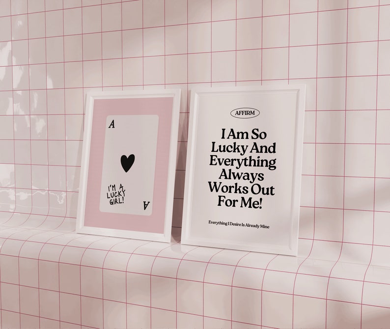 Puede incluir: Dos impresiones enmarcadas, una con fondo rosa y una carta de juego de as de corazones negra con el texto "I'm a lucky girl!" y la otra con fondo blanco y el texto "AFFIRM I Am So Lucky And Everything Always Works Out For Me! Everything I Desire Is Already Mine."