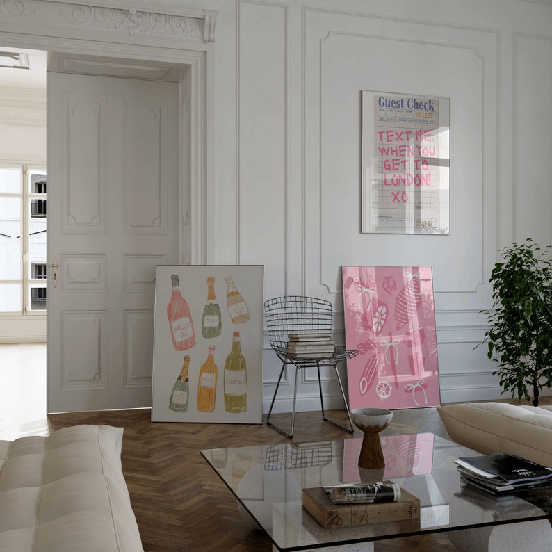 May include: A living room interior with a white wall, a white door, a glass coffee table, a white couch, a metal chair, and two framed prints. One print features a pink and white illustration of a fish and the other print features a pink and white illustration of bottles of wine. The text on the wall reads "Guest Check" and "Text me when you get to London! XO!"