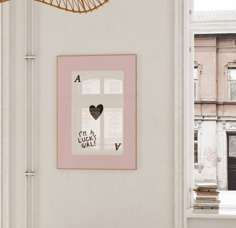 Puede incluir: Una impresi&oacute;n enmarcada rosa de una carta de juego de as de corazones con un coraz&oacute;n negro en el centro y el texto "I'm a lucky girl!" escrito en ella.