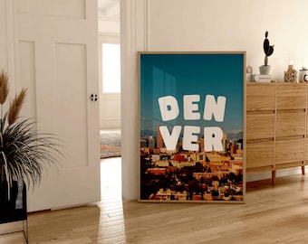 May include: A framed print of a cityscape with the word "DENVER" in large white letters against a blue sky. The city is in the foreground and the sky is in the background.