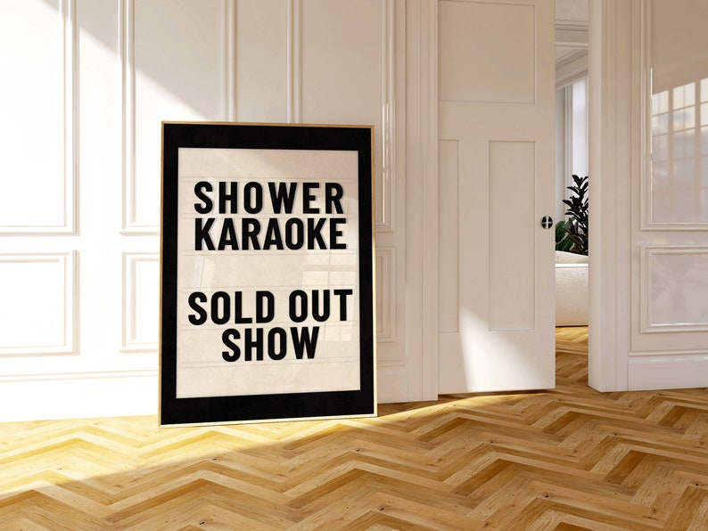 以下が含まれることがあります： 白い背景に黒い文字で「Shower Karaoke Sold Out Show」と書かれた、黒い額縁のポスター。ポスターは白い壁にかけられており、部屋には木製の床がある。