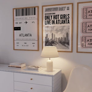 May include: A framed print featuring a black and white image of a boarding pass for a flight to Atlanta. The print is hanging on a white wall alongside a framed print of a newspaper article with the headline "Only Hot Girls Live in Atlanta".