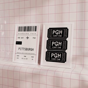 Może przedstawiać: Biały obraz w ramie z trzema czarno-białymi biletami z napisem "PGH Admit One" oraz biały obraz w ramie z kartą pokładową z napisem "Boarding Pass One Way PGH First Class Pittsburgh Gate 422 Time 01:11 EST Seat 18" na tle r&oacute;żowo-białych płytek.