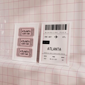 May include: A white framed picture with three tickets for admission to Atlanta. The tickets are printed with the text "Atlanta Admit One" and the number "552142".  A white framed picture with a boarding pass for a one-way flight to Atlanta. The boarding pass is printed with the text "Boarding Pass", "One Way", "ATL", "First Class", "Atlanta", "Gate A22", "Time 01:11 EST", and "Seat 18".