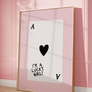 Puede incluir: Una impresi&oacute;n enmarcada de una carta de juego con un coraz&oacute;n negro y el texto "I'm a lucky girl!" sobre un fondo rosa.