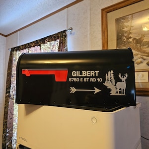May include: Black mailbox with a red flag and white lettering that reads "GILBERT 5750 E ST RD 10". A white arrow points to the right. A white silhouette of a deer and trees is on the right side of the mailbox.
