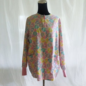 Puede incluir: Una bata de cirujano rosa con un estampado de huevos de Pascua coloridos. La bata tiene un cierre de botones en la parte delantera y mangas largas con puños rosas.