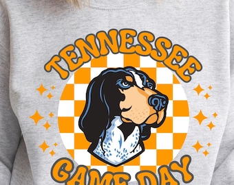Puede incluir: Una sudadera gris con un diseño gráfico que presenta un perro de caza negro y blanco con un collar azul y blanco. El gráfico está rodeado por un círculo a cuadros naranja y blanco con el texto "Tennessee Game Day" en naranja.