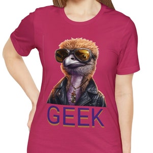 Op de afbeelding: Een roze T-shirt met een afbeelding van een struisvogel die een zonnebril en een leren jack draagt. De tekst "GEEK" is in blauw en oranje op het T-shirt gedrukt.