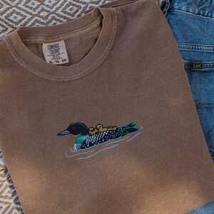 May include: A brown Comfort Colors t-shirt featuring an embroidered loon and chicks design, alongside blue denim jeans. The shirt has a tag that reads XL-TG-EG. The image shows a casual outfit.