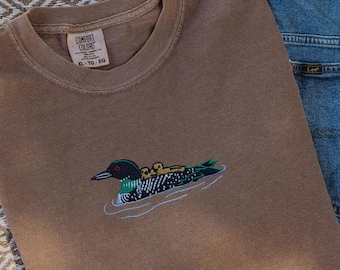 May include: A brown Comfort Colors t-shirt with an embroidered design of a loon and its chicks swimming in water. The shirt is paired with blue denim jeans. The shirt has a tag that reads XL-TG-EG.