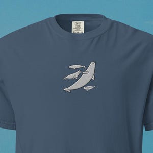 Könnte beinhalten: Blaues T-Shirt mit einer weißen Illustration eines Wals und zwei kleineren Walen. Das T-Shirt hat einen Rundhalsausschnitt. Das Bild ist vor einem Strand- und Ozeanhintergrund platziert.