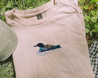 May include: A beige t-shirt with a detailed embroidered design of a loon swimming in water. A green baseball cap with a loon design and a green and white checkered fabric are also visible.