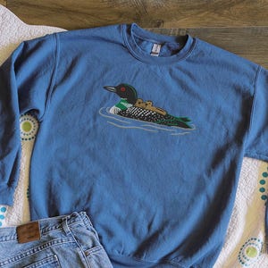 May include: A blue sweatshirt featuring an embroidered design of a loon with two chicks swimming in water. The sweatshirt is paired with blue jeans, a white fanny pack, and a water bottle. The sweatshirt is a crew neck style.