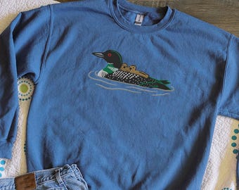 Puede incluir: Sudadera azul con un diseño bordado de un somorgujo con dos crías nadando en el agua. La sudadera se combina con vaqueros azules, una riñonera blanca y una botella de agua. El diseño del somorgujo es en tonos verdes, negros y marrones.