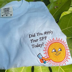 May include: Light blue t-shirt with the text "Did You Apply Your SPF Today?" and a smiling sun graphic holding sunscreen. The shirt is a Comfort Colors brand.