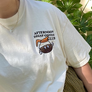 May include: Cream-colored t-shirt with the text "Afternoon Decaf Coffee Club" and a cartoon coffee pot graphic. The shirt is made of a soft material and is suitable for casual wear. The shirt is paired with a yellow and black plaid skirt.