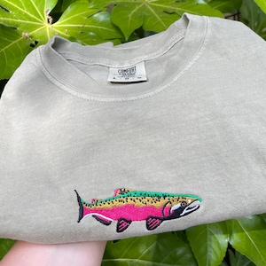 May include: A sage green sweatshirt with an embroidered rainbow trout design. The fish features pink, yellow, and green details. The sweatshirt is folded and set against a backdrop of large green leaves.