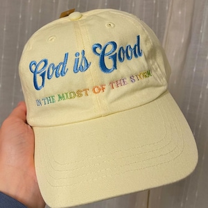 Puede incluir: Gorra de béisbol de color amarillo claro con la frase "God is Good" en escritura azul. Debajo, en colores arcoíris, el texto "IN THE MIDST OF THE STORM". La gorra tiene una visera curva y una correa de tela.