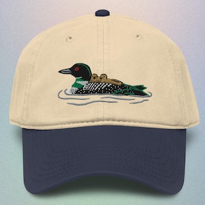 May include: A baseball cap with a cream-colored crown and a navy blue brim. The cap features an embroidered design of a loon with its chicks swimming on water. The loon has a black head, red eye, and green and black body.