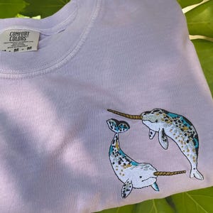 May include: A light purple sweatshirt with embroidered narwhals. The narwhals have blue, white, and gold accents. The sweatshirt has a crew neck and the brand "Comfort Colors" is visible on the tag.