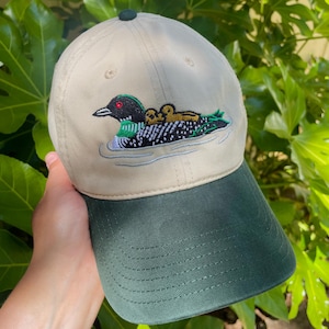 Könnte beinhalten: Eine beige Baseballkappe mit dunkelgrünem Schirm und einem dunkelgrünen Knopf an der Oberseite. Auf der Vorderseite befindet sich eine Stickerei eines schwarz-weißen Eistauchers mit rotem Auge, der zwei kleine braune Küken auf dem Rücken trägt.