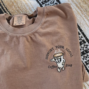May include: Brown t-shirt with an embroidered design of a coffee cup character with the text "Support Your Local Coffee Shop".